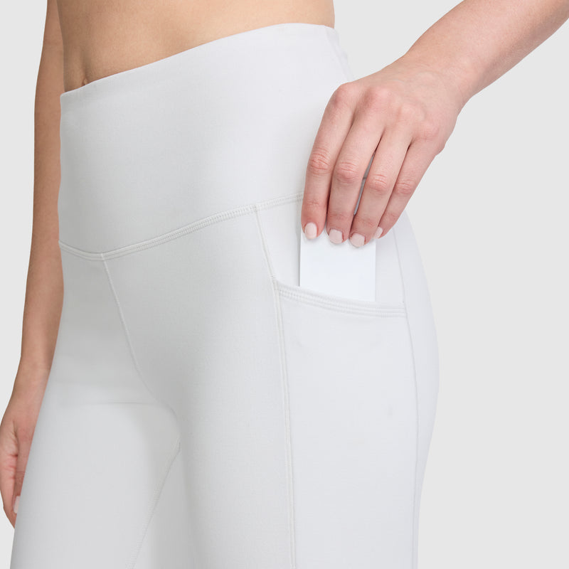 A person wearing Champion’s C Logo high-waisted white leggings with handy side pockets slides a white card or phone into one of the pockets. Only the lower torso and hand are visible against a plain light background, drawing attention to the smooth peached fabric.