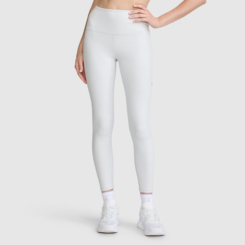 A person models the Champion C Logo Leggings, featuring a high-waisted white design with side pockets. Paired with white sneakers and socks, they stand against a plain light gray background, left hand resting on their hip and only the lower half of their body shown.
