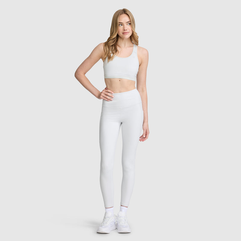 A woman with light hair and a neutral expression stands against a plain background wearing Champion’s C Logo high-waisted leggings, crafted from peached fabric and featuring subtle side pockets for added style and function. She pairs the leggings with a white sports bra and white sneakers, resting one hand on her hip.