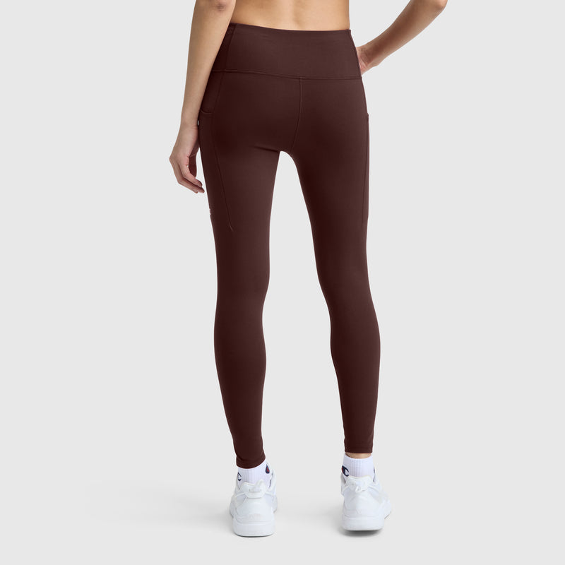 A person wearing Champion C Logo high-waisted, dark brown silky soft leggings and white sneakers stands with their back to the camera against a plain light gray background.