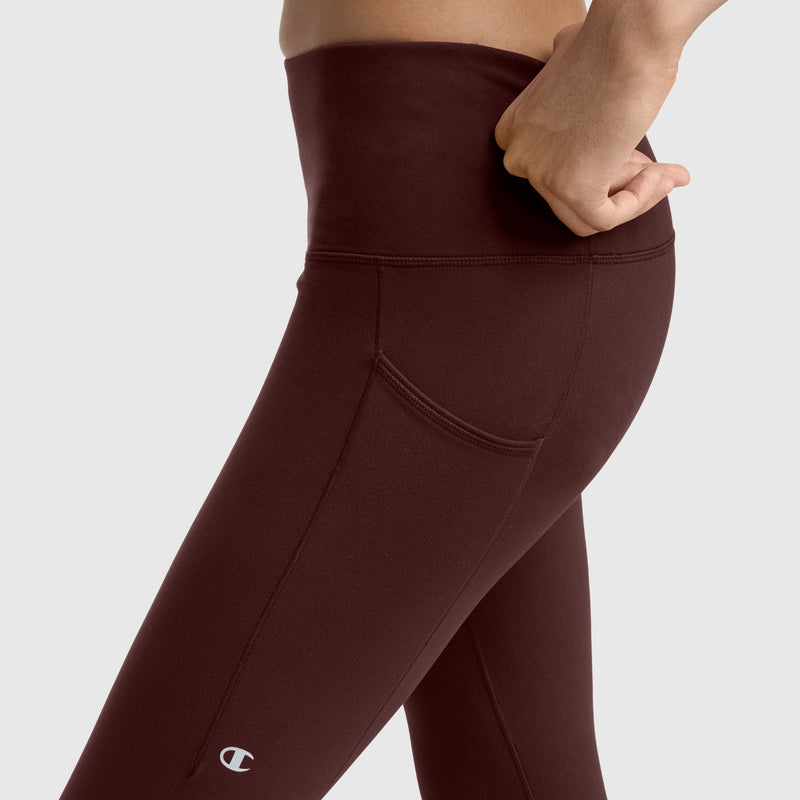 A person wearing Champion C Logo high-waisted maroon leggings with pockets is standing, with one hand resting on their hip. The Champion logo is visible near the knee.
