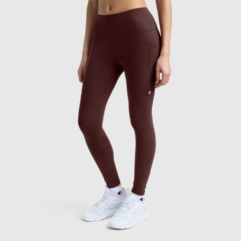 A person wearing Champion’s C Logo high-waisted, silky soft leggings in maroon paired with white sneakers stands against a plain light background, arms relaxed at their sides.