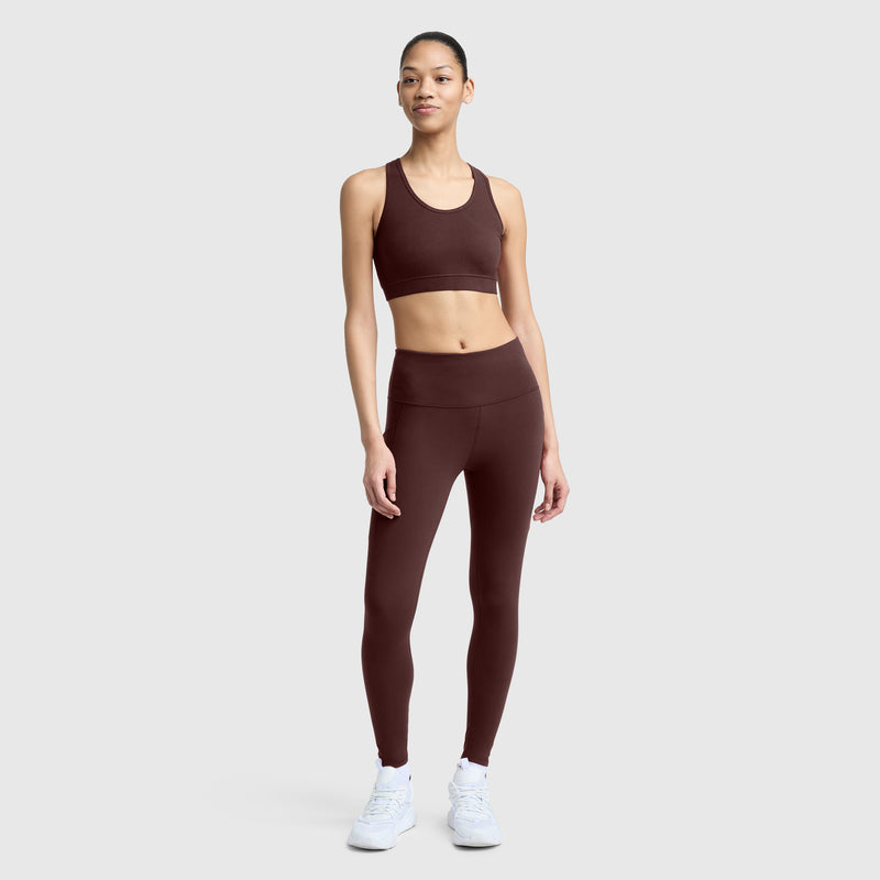 A woman stands against a plain background wearing the Champion C Logo leggings paired with a matching maroon sports bra and white sneakers. She faces forward with a neutral expression and relaxed posture, highlighting the silky soft leggings.