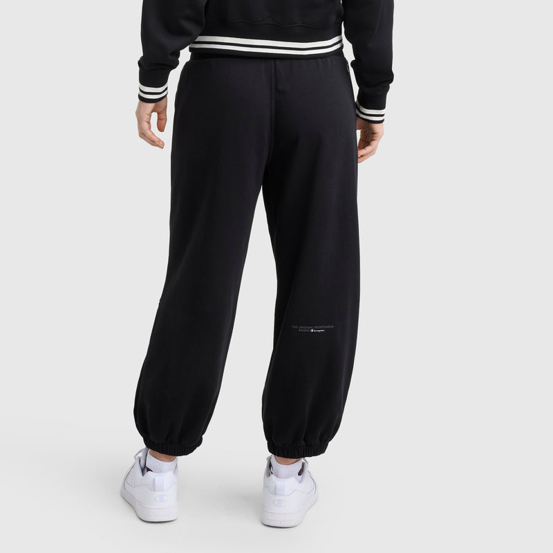 A person wearing Champion Tackle Snap Front Voluminous Pant in black and white sneakers stands with their back to a plain light gray background. The pants have elastic cuffs and small white text near the right calf.