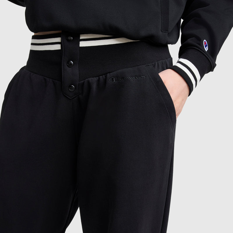 Someone wearing Champion's Tackle Snap Front Voluminous Pant in black, featuring an embroidered logo, paired with a black jacket with white striped cuffs and waistband, shown waist down with one hand in the pocket.