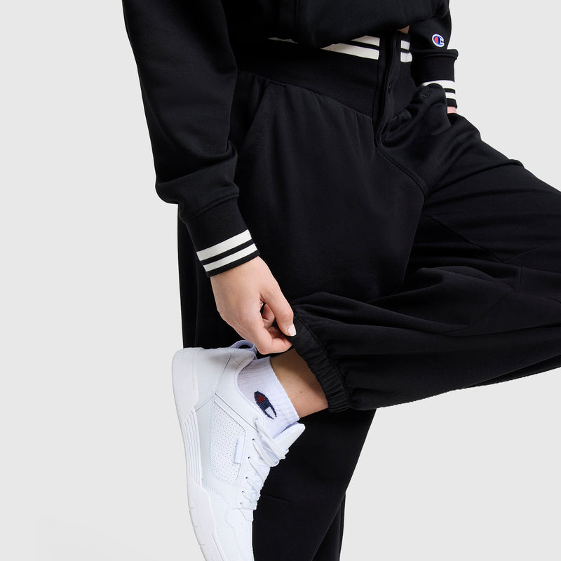 A person wears the Champion Tackle Snap Front Voluminous Pant with a matching black jacket featuring white striped cuffs, lifting one leg to show white socks and sneakers. Only the lower half is visible, evoking a vintage athletic vibe.