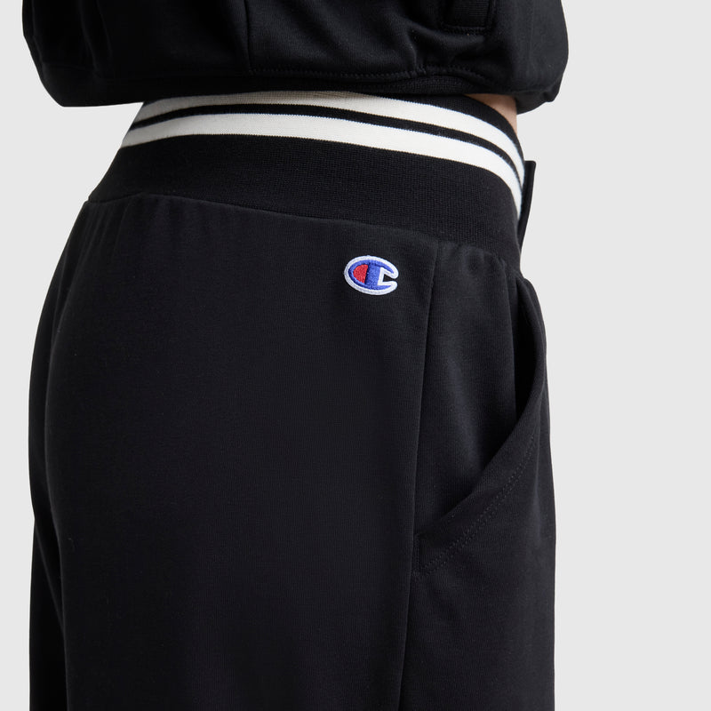 Close-up of a person wearing Champion's Tackle Snap Front Voluminous Pant, featuring soft French terry fabric, a striped waistband, and the Champion logo on the hip for a vintage athletic vibe against a plain light background.