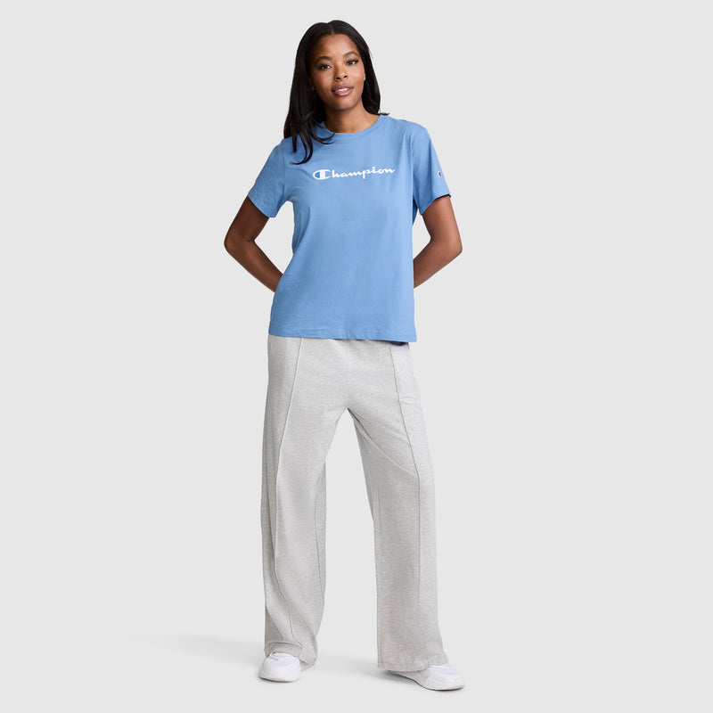 A woman stands against a plain background, wearing a blue Champion t-shirt, light gray Champion Open Bottom Pant crafted from soft stretch jersey, and white sneakers, with her hands resting on her hips.