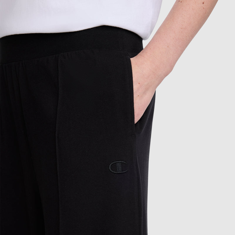 A person is shown wearing black Champion Open Bottom Pant with a hand in the pocket. The image features only the lower torso and part of the arm, and the individual is also dressed in a white shirt.