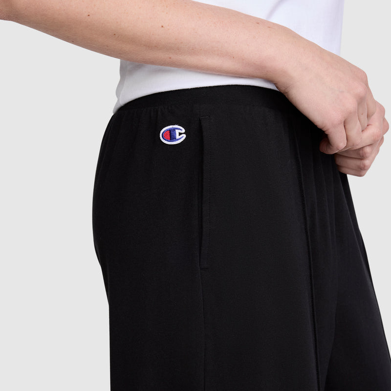 A person is seen wearing Champion Open Bottom Pants in black stretch jersey with the signature logo near the pocket, styled with a white shirt. The image focuses on the midsection, arm, and hand against a plain background, showcasing the athletic design of these Champion pants.