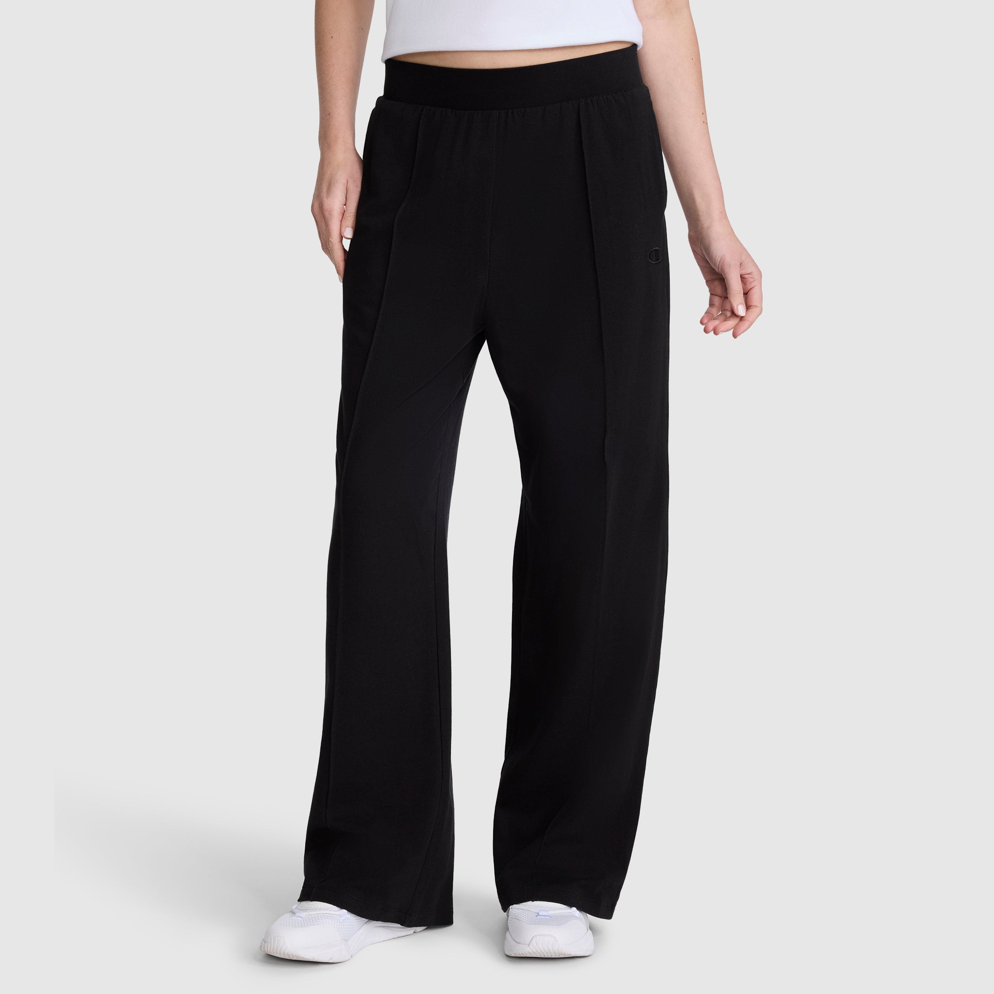 A person wearing Champion's Open Bottom Pant in black stretch jersey, paired with a white sleeveless top and white sneakers, stands against a plain light gray background. Only the lower half of the body is visible.