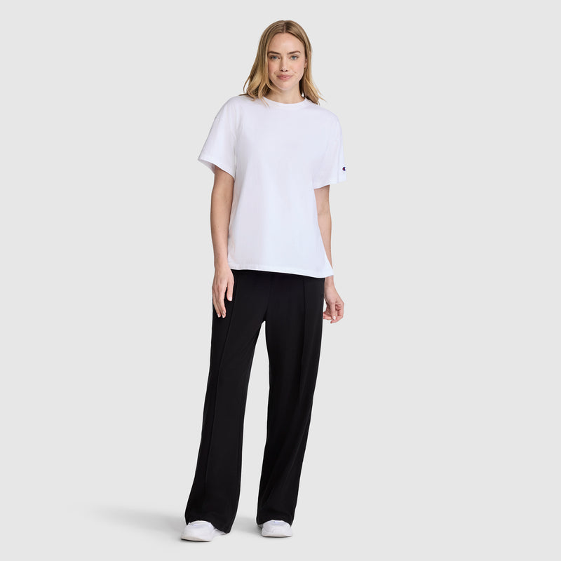 A woman stands against a plain background, wearing a loose white T-shirt, Champion Open Bottom Pant in black, and white sneakers. She has blonde hair, a slight smile, and her arms are relaxed by her sides.