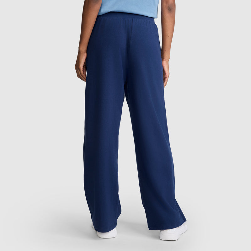 A person stands facing away against a plain background, wearing Champion Open Bottom Pants in a loose-fitting dark blue athletic style with a light blue shirt and white shoes, displaying the back of the outfit.