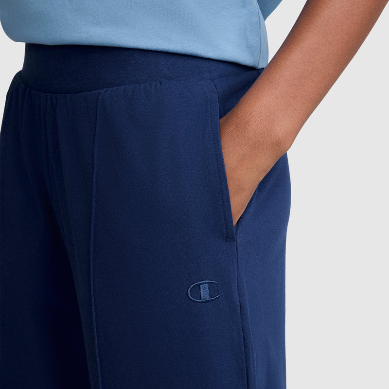 A person wearing the Champion Open Bottom Pant in navy blue, styled like stretch jersey pants with an embroidered logo on the thigh and a hand in the pocket. The person is also wearing a light blue shirt.