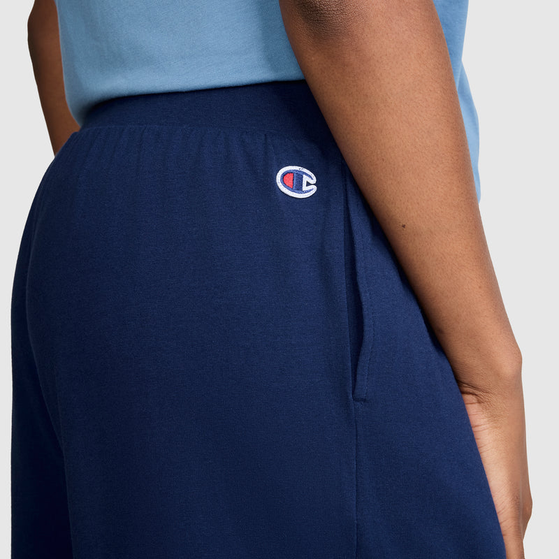 A person is shown from the side wearing the Champion Open Bottom Pant in navy blue with a light blue shirt, highlighting the Champion logo near the waistband. These pants provide athletic style along with comfort and versatile everyday wear.