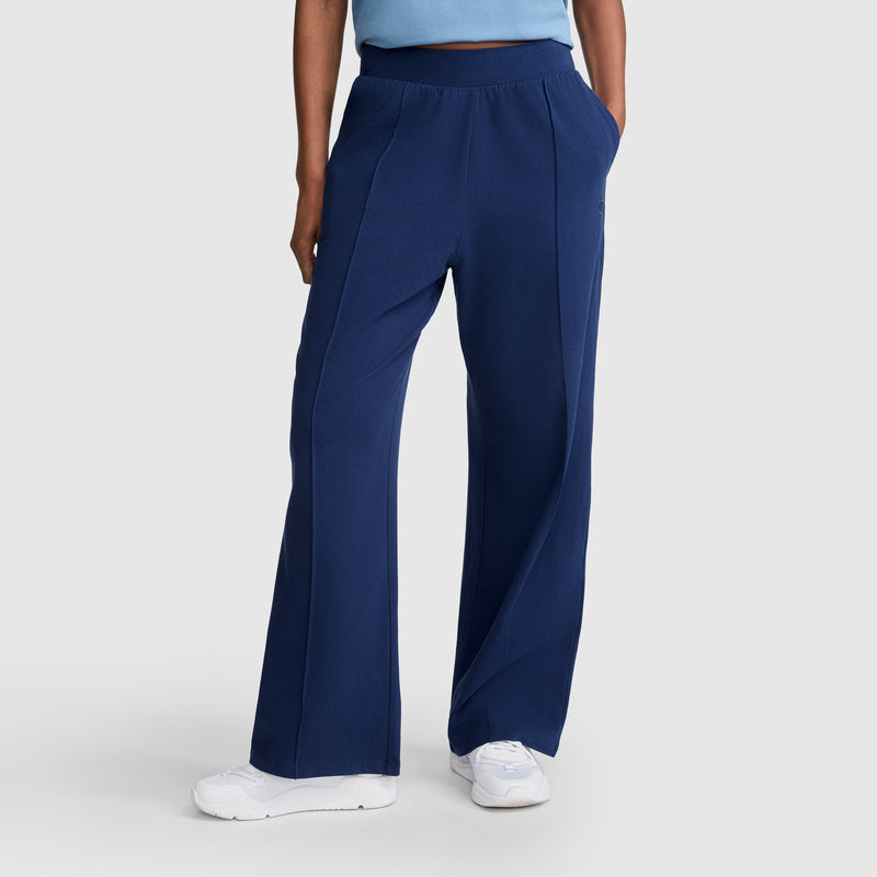 A person wearing Champion Open Bottom Pant in navy blue stretch jersey, paired with a light blue top and white sneakers, stands against a light grey background.