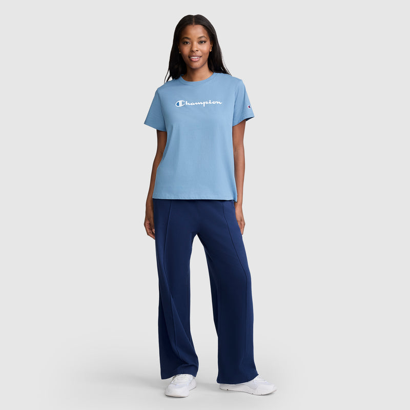 A woman stands and smiles, wearing a light blue Champion T-shirt, dark blue Champion Open Bottom Pant, and white sneakers against a plain light gray background.