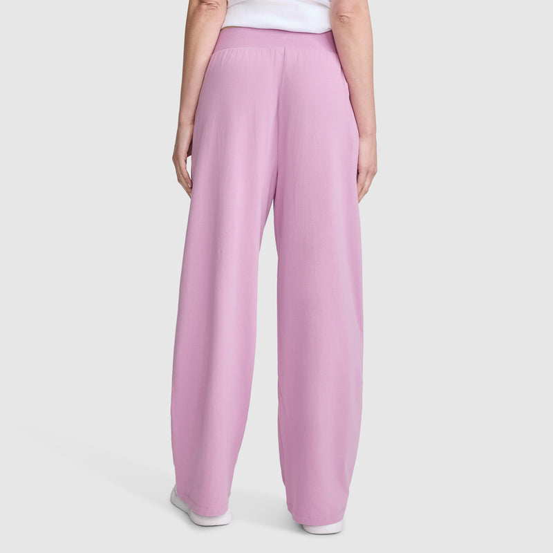 A person is shown from the back standing against a plain light background, wearing Champion Open Bottom Pant in light pink—a loose, wide-leg stretch jersey style—paired with a white top.