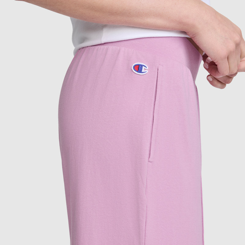 A person is shown from the waist down in a side view, wearing light pink Champion Open Bottom Pant and a white top, with one hand resting near their hip.