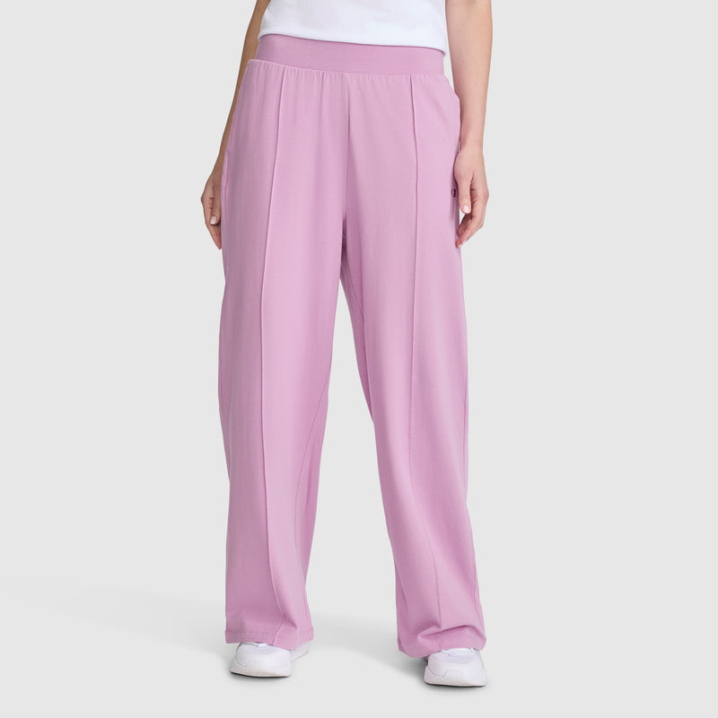 A person wearing Champion Open Bottom Pant in high-waisted pink stretch jersey, paired with a white shirt and white sneakers, stands against a plain light gray background.