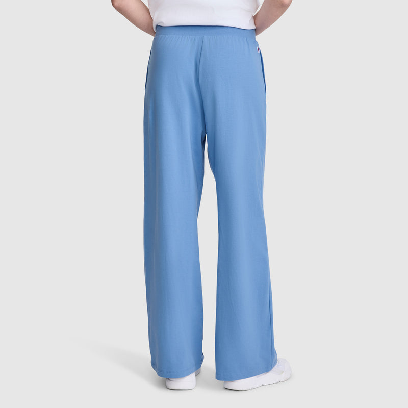 A person wearing Champion Open Bottom Pant athletic pants in light blue, featuring a wide-leg silhouette and crafted from soft stretch jersey fabric, paired with a white t-shirt. The back view is set against a plain light gray background, with the small Champion logo visible on the upper right waistband.
