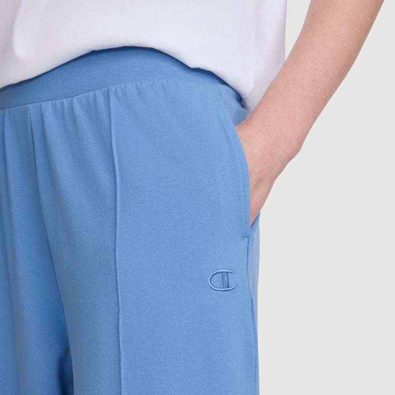 A person is wearing light blue Champion Open Bottom Pant stretch jersey athletic pants, featuring the Champion logo and with one hand in the pocket. The pants are paired with a white shirt, and only the lower torso and part of one arm are visible.