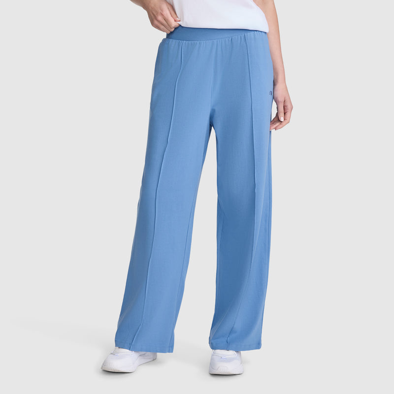 A person wearing Champion’s Open Bottom Pant—loose-fitting, light blue athletic pants made from comfortable stretch jersey with an elastic waistband—paired with white sneakers and a white top, stands against a plain light gray background.