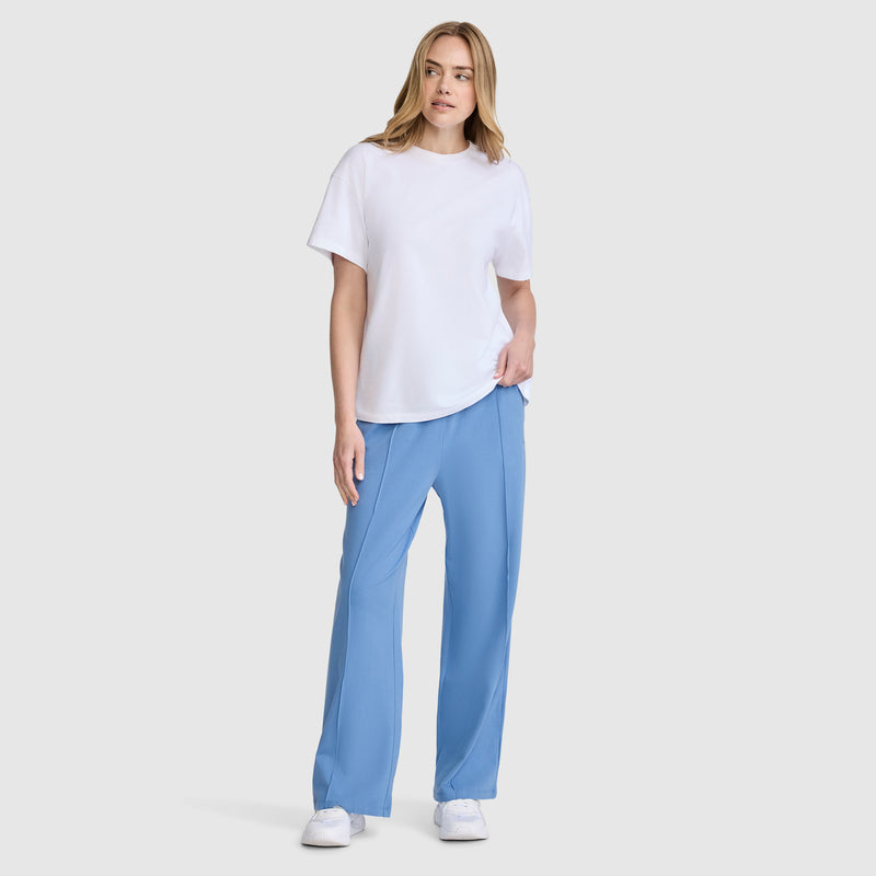 A woman with light hair poses casually against a plain background, wearing a white short-sleeve t-shirt, the Champion Open Bottom Pant in light blue made from stretch jersey, and white sneakers. She has one hand in her pocket for a relaxed look.