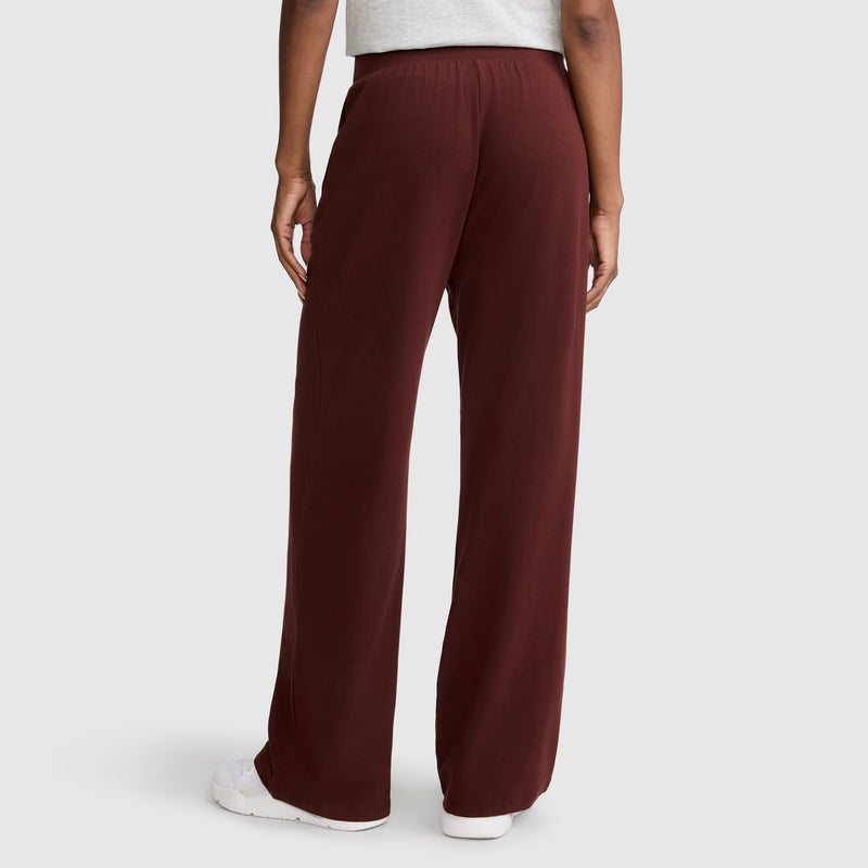 Seen from behind, a person wears Champion’s Open Bottom Pant for women in maroon stretch jersey fabric, paired with a light gray shirt and white sneakers, creating an effortless athletic look against a simple light background.