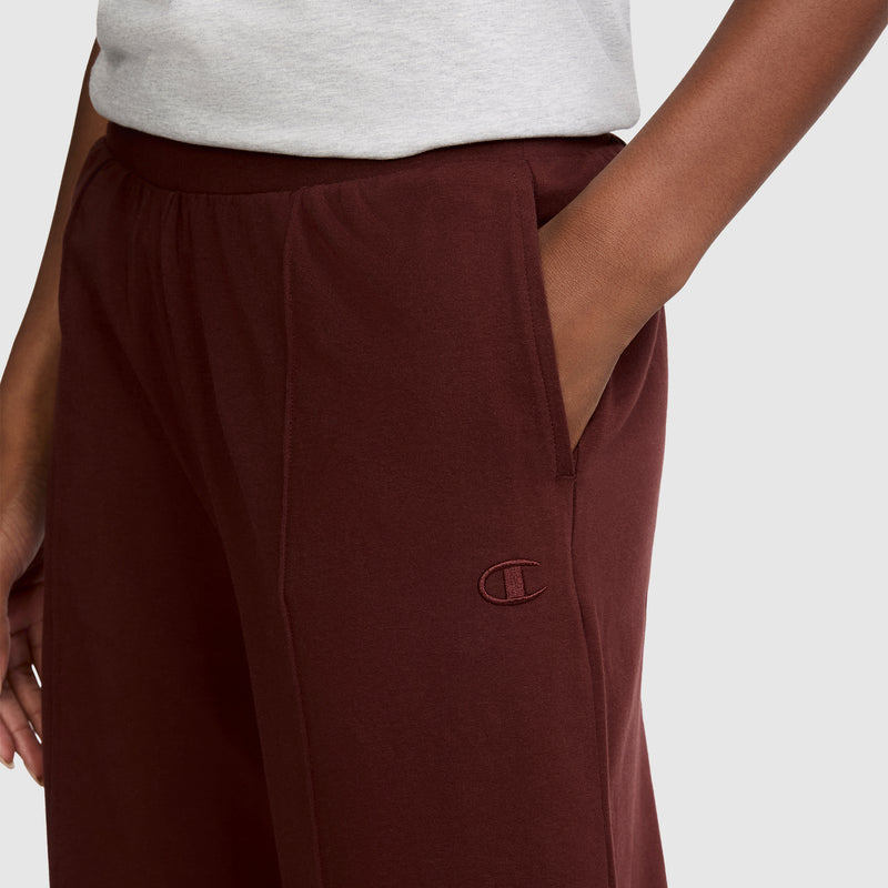 A person wearing the Champion Open Bottom Pant in maroon, with a hand placed in the pocket, paired with a light gray shirt. The stretch jersey pants showcase an embroidered Champion logo near the pocket, enhancing their athletic look.
