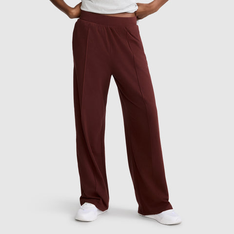 A person wears Champion’s Open Bottom Pant—a high-waisted, wide-leg maroon stretch jersey style with an athletic silhouette—paired with a tucked white shirt and white sneakers, standing against a plain light gray background. Only the lower body is visible.