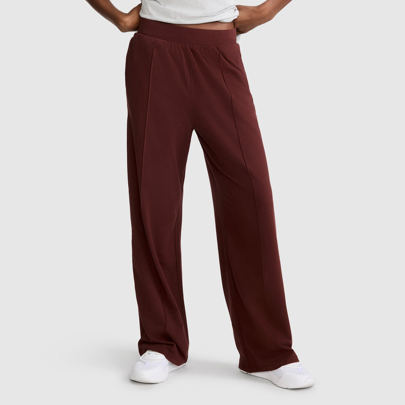 A person wears Champion’s Open Bottom Pant—a high-waisted, wide-leg maroon stretch jersey style with an athletic silhouette—paired with a tucked white shirt and white sneakers, standing against a plain light gray background. Only the lower body is visible.