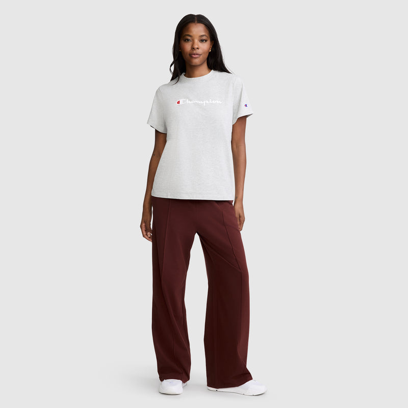 A woman stands facing forward against a plain light gray background, wearing a light gray Champion T-shirt, Champion Open Bottom Pant in maroon, and white sneakers.