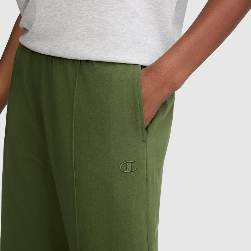 Someone is wearing olive green Champion Open Bottom Pants, featuring a side pocket and a small C logo, paired with a light gray shirt. Only the lower torso and arm are visible.