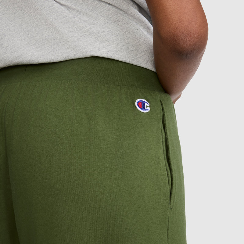 A person wearing Champion Open Bottom Pants in olive green, featuring the signature logo on the hip, paired with a light gray shirt. Only part of the torso and arm are visible, emphasizing the comfort and style of these Champion pants.