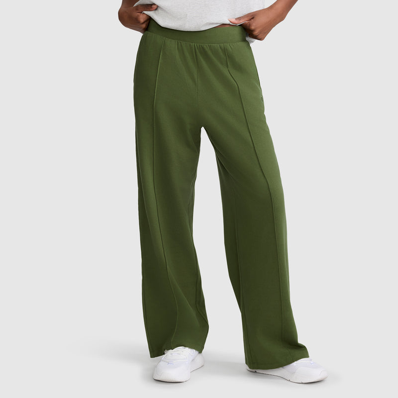 A person is shown from the waist down, wearing Champion Open Bottom Pants in loose-fitting olive green stretch jersey with hands in pockets, paired with white sneakers, standing against a plain background.