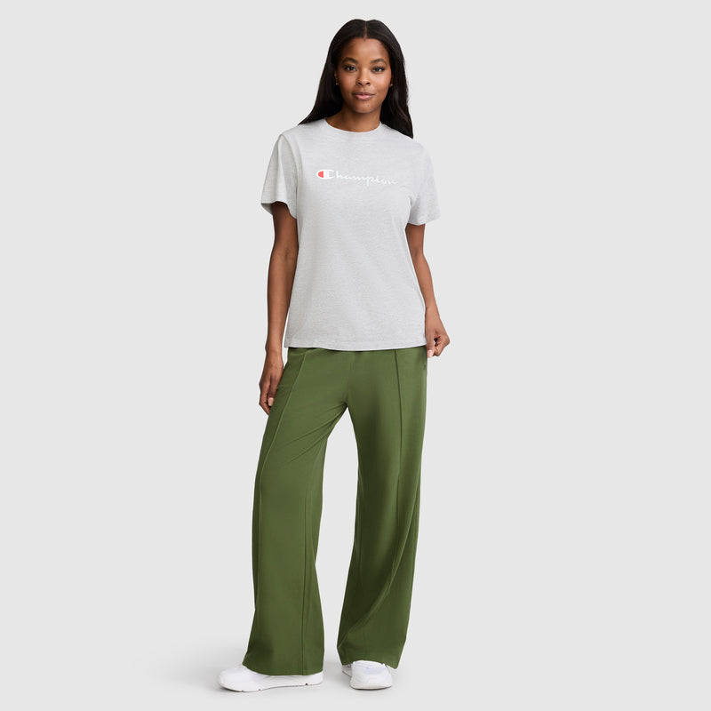 A woman stands smiling against a plain light gray background, wearing a light gray Champion t-shirt, white sneakers, and the Champion Open Bottom Pant—a loose-fit style in olive green stretch jersey.