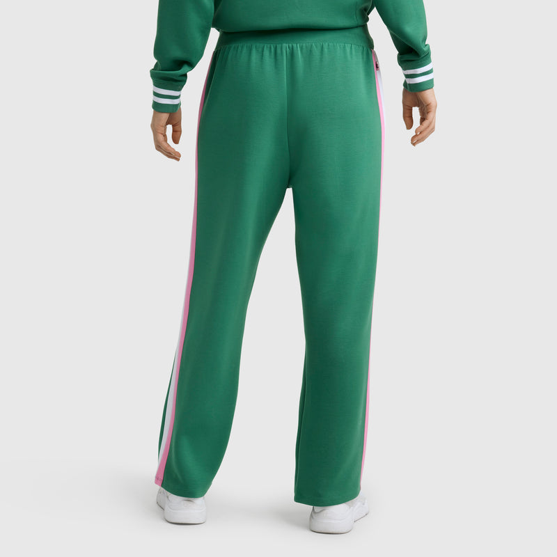 A person wearing Champion's Tennis Court Wide Leg Pant, C Logo in green with pink and white side stripes, paired with a matching top and white sneakers, stands with their back to the camera in sporty style.