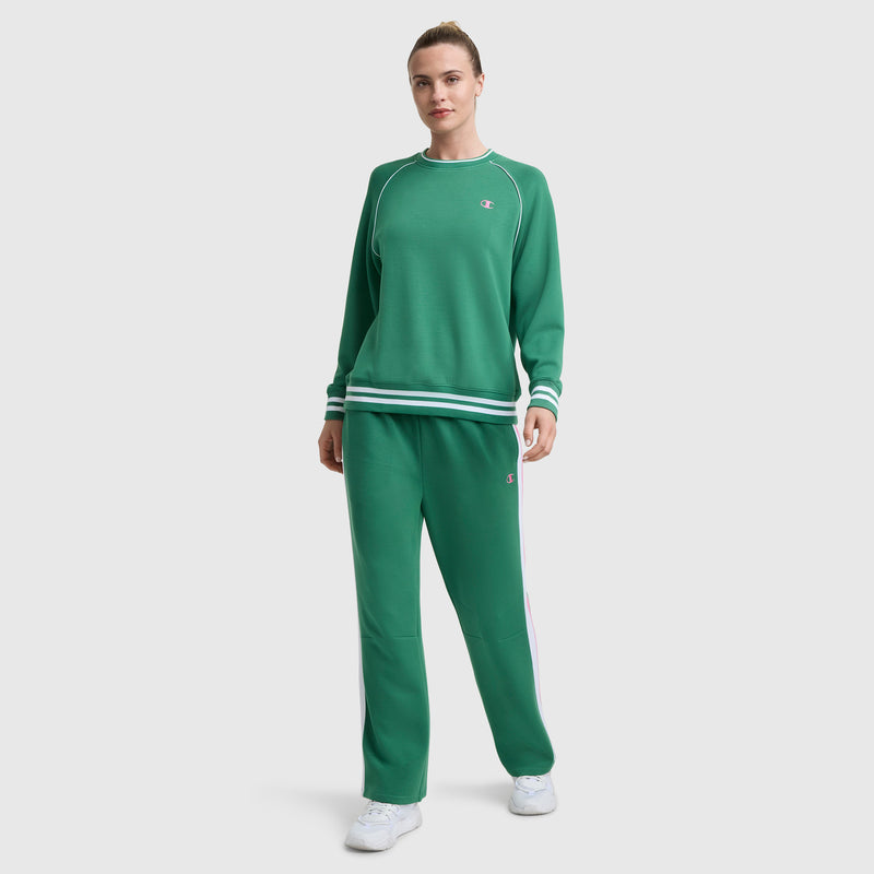 A person stands facing forward in Champion’s Tennis Court Wide Leg Pant featuring the C Logo and white and green striped cuffs and waistband, crafted from soft scuba fabric, paired with white sneakers. The plain background is light-colored.