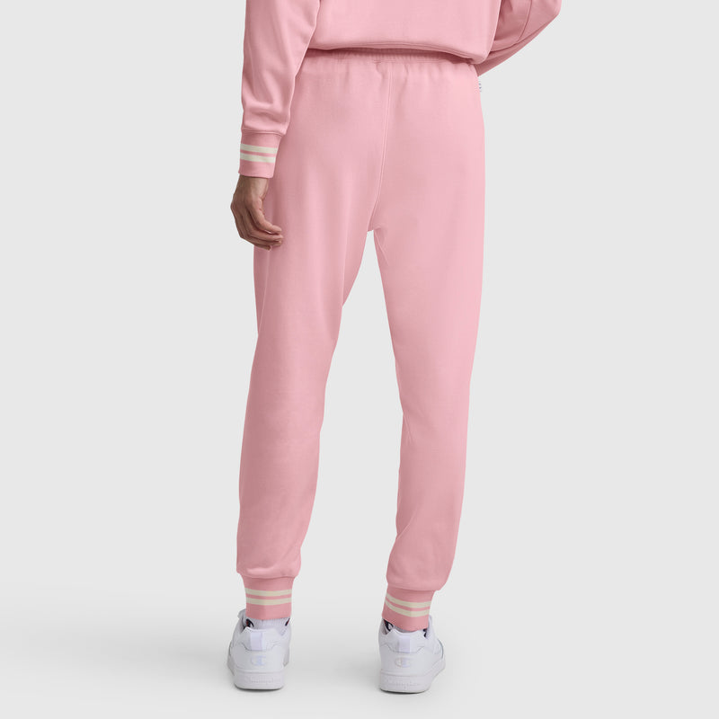 Seen from behind, a person wears Champion Pitcher Pant, C Logo in light pink French terry with elastic waistband and cuffs, paired with a matching pink and white sweatshirt and white sneakers on a white background.