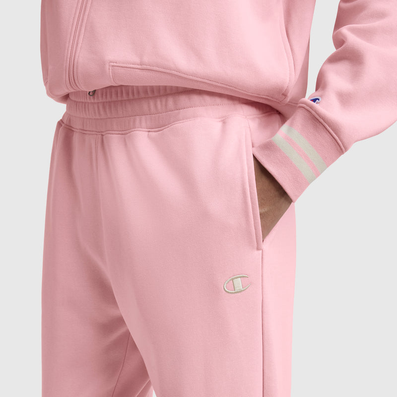 Someone wears Champion’s Pitcher Pant with the C Logo in light pink, featuring white striped cuffs and their hand in the pocket. Only their lower torso and arm are visible.
