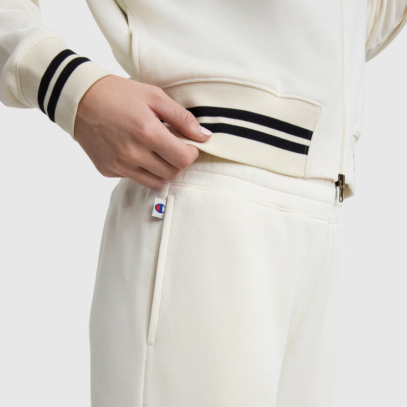 A person in off-white Champion Pitcher Pants with a C logo tag near the pocket adjusts their jacket. The French terry joggers have black and white striped cuffs. The background is plain white.