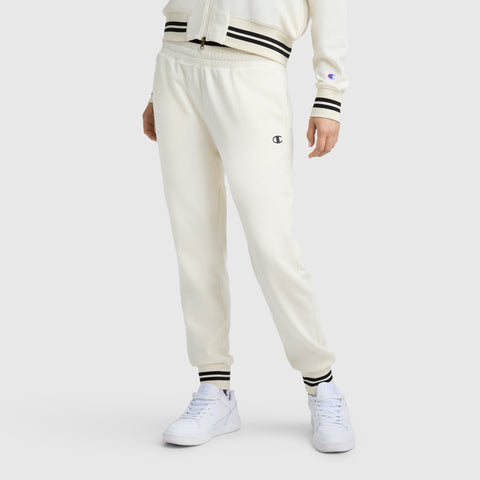 A person wears Champion Pitcher Pant joggers with black and white striped cuffs and waistband, featuring the C Logo on the left thigh. Paired with white sneakers; upper body is partially visible.