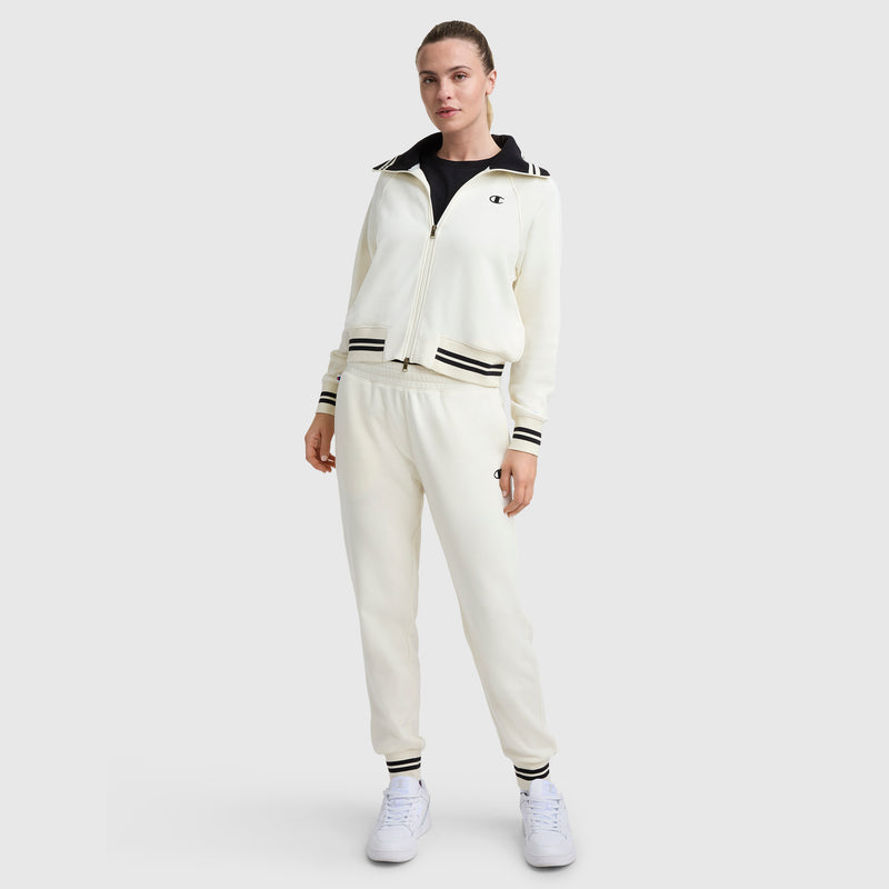 A woman wears Champion's Pitcher Pant, C Logo—a cream sport-luxe tracksuit with black stripes on collar, cuffs, and waistband—paired with white sneakers, creating a chic athletic look against a light gray background.