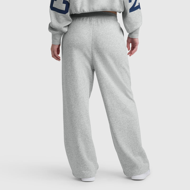 A person stands facing away from the camera in light gray Champion Tackle Open Bottom Pant, C Logo, paired with a matching sweatshirt featuring navy numbers on the sleeves, against a plain white background.