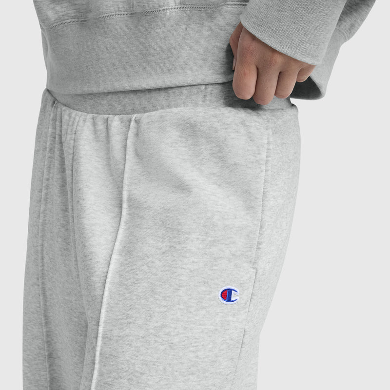 A person wears Champion’s Tackle Open Bottom Pant, C Logo in light gray French terry with a matching sweatshirt, hand in pocket. The Champion logo appears on the wide leg of the pant.