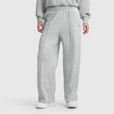 A person wears Champion’s Tackle Open Bottom Pant with C Logo in light gray, featuring a drawstring waist and front seams, paired with a matching sweatshirt and white sneakers against a plain white background.