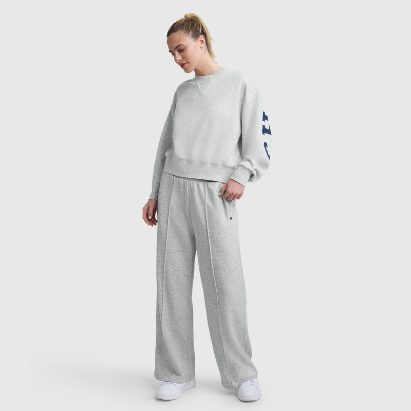 A woman, head slightly tilted down and one hand in her pocket, stands against a white background wearing Champion’s Tackle Open Bottom Pant with C Logo in light gray French terry and a matching loose sweatshirt, paired with white sneakers.