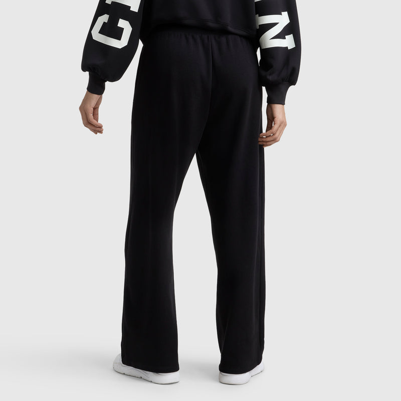 A person faces away, showcasing Champion's Tackle Open Bottom Pant and a black sweatshirt with white sleeve lettering, paired with white sneakers against a plain background.