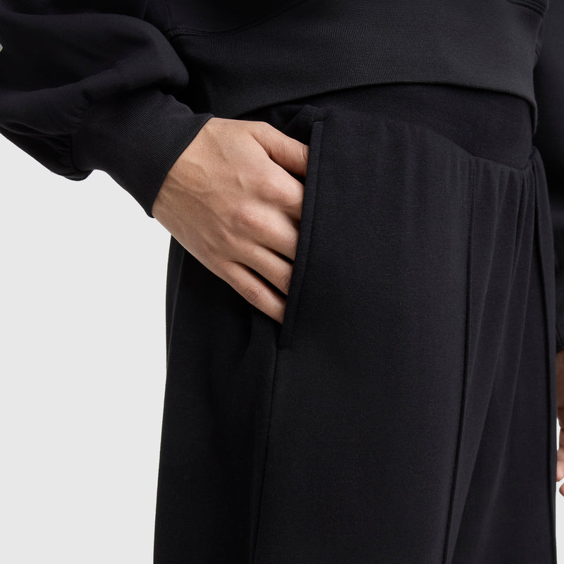 A person is wearing Champion’s Tackle Open Bottom Pant, C Logo in black with a matching black long-sleeve top, their right hand in the pant pocket against a plain light background.